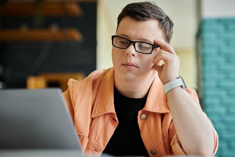 Young man with glasses struggling to see laptop and adjusting his glasses.